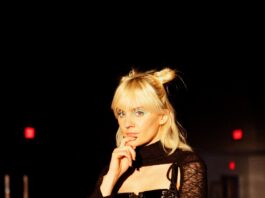 Liza Anne is white with light blonde hair and bangs. They are wearing an all-black outfit with a skirt and fishnet tights. They are standing and holding a blue and white electric guitar.