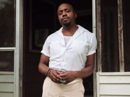 Durand Jones stands in a doorway. He is wearing a white shirt and light colored pants. He is also wearing bracelets and a ring.
