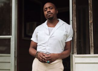 Durand Jones stands in a doorway. He is wearing a white shirt and light colored pants. He is also wearing bracelets and a ring.