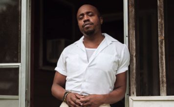 Durand Jones “See It Through” – Dan’s DJ Pick of the Week Durand Jones stands in a doorway. He is wearing a white shirt and light colored pants. He is also wearing bracelets and a ring.